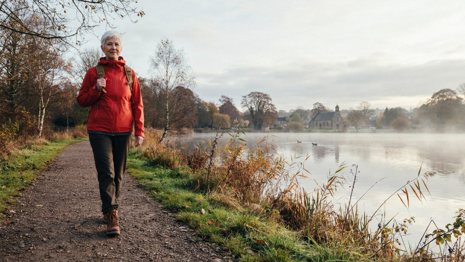 Older adult walking beside Duddingston Loch in Edinburgh on a still late autumn morning, the age group offered the 2026/27 COVID-19 booster