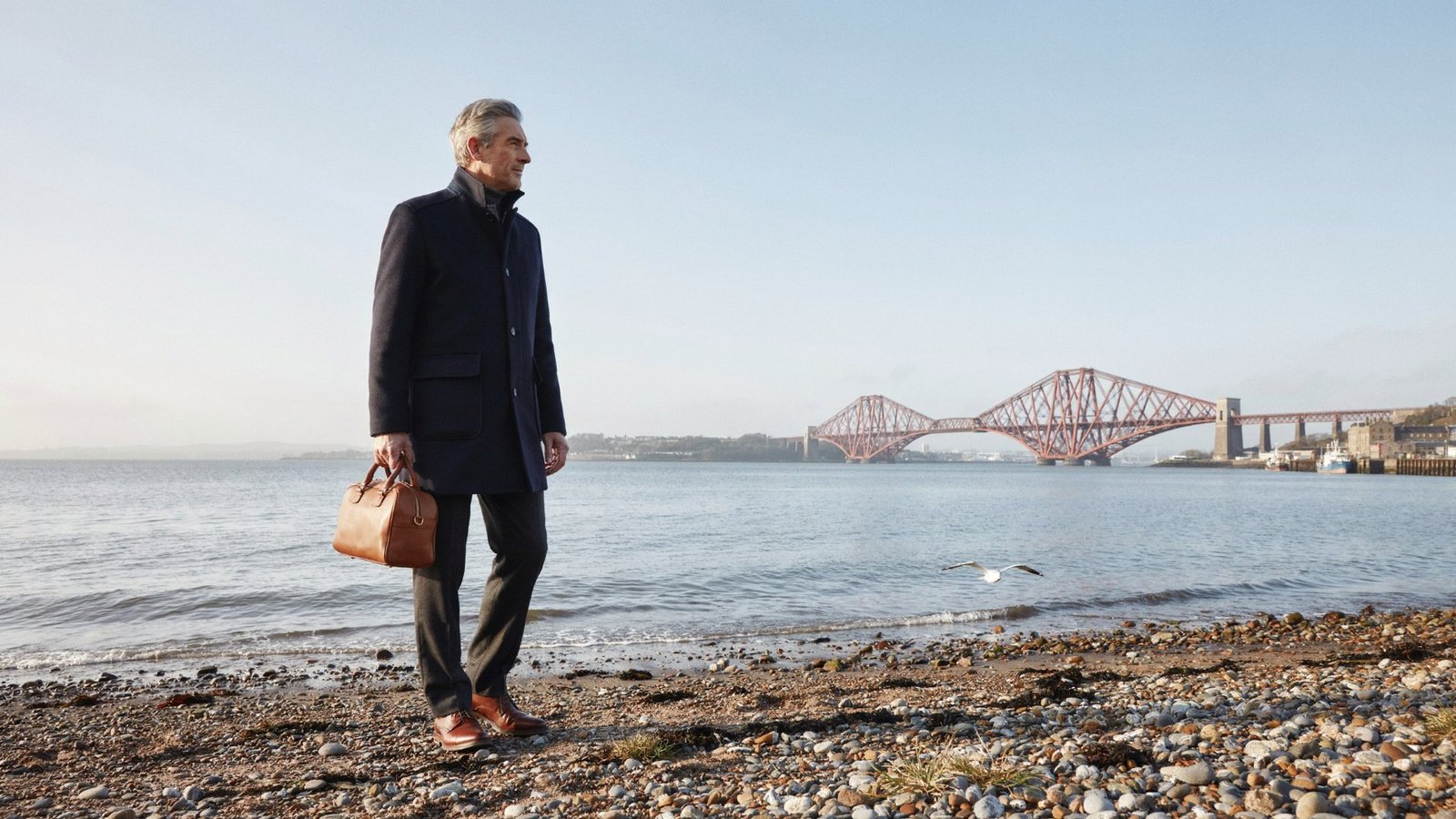 Adult with a small travel bag on the shore at South Queensferry, with the Forth Bridge behind, on a bright winter morning before a trip, protected for the season